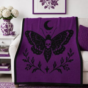 May include: A purple knit throw blanket featuring a black graphic design of a moth with a skull on its thorax, positioned beneath a crescent moon and above two leafy branches with a small four-pointed star centred at the bottom edge.