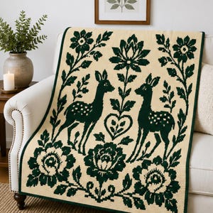 May include: A rectangular knit throw blanket featuring a cream-coloured background with a dark green pattern. The design displays two deer facing each other, surrounded by large floral motifs, leafy vines, and a central heart shape in a pixelated style.