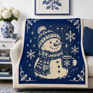 Op de afbeelding: Een rechthoekige plaid met een donkerblauwe achtergrond en een crèmekleurig gebreid patroon van een sneeuwpop met een muts met pompon en een sjaal, die een mok vasthoudt. Het ontwerp bevat sneeuwvlokken en sterren binnen een decoratieve rand.