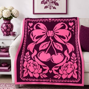 May include: A rectangular knit throw blanket featuring a deep purple background with a vibrant pink graphic design. The pattern displays a large central bow tied above two cherries, flanked by roses at the bottom and small hearts at the top.