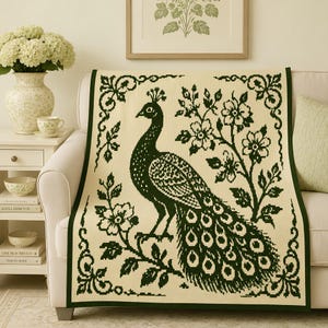 Puede incluir: Una manta de color crema con un diseño tejido en verde oscuro de un pavo real de pie entre ramas florales. La manta está bordeada por un marco decorativo de volutas, que muestra un patrón detallado de hojas y flores.