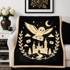 May include: A black throw blanket with a beige border featuring a pixelated graphic of a flying owl carrying a letter above a castle, surrounded by clouds, stars, a crescent moon, and a decorative wreath of leaves on a textured fabric surface.
