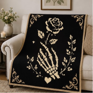 Pode incluir: Uma manta retangular preta com um design bege pixelizado de uma mão de esqueleto segurando uma rosa. A manta inclui ornamentos decorativos nos cantos e um pequeno coração na parte inferior, tudo contornado por uma borda bege sólida.