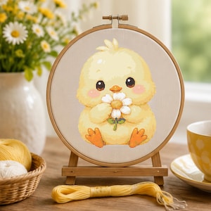 May include: A round wooden embroidery hoop containing a cross-stitch pattern of a fluffy yellow chick with large dark eyes and pink cheeks, holding a white daisy flower with a yellow center. The hoop is mounted on a small wooden display stand.
