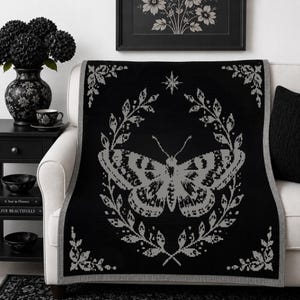May include: A black throw blanket featuring a large, centered grey moth graphic enclosed within a detailed botanical wreath. The design includes a small star icon above the moth and decorative floral corner accents, finished with a grey border.