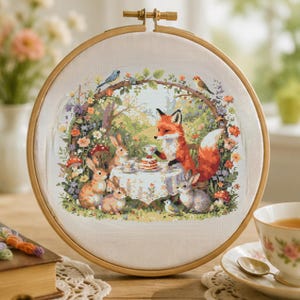 May include: A circular wooden embroidery hoop framing a cross-stitch design of a fox and three rabbits having a tea party. The scene features a table with a cake, surrounded by colourful flowers, mushrooms, and small birds in a garden setting.