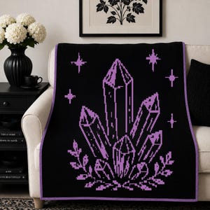 May include: A black throw blanket featuring a purple pixel art design of a crystal cluster surrounded by small four-pointed stars and leafy sprigs. The blanket has a solid purple border trim that frames the rectangular fabric piece.