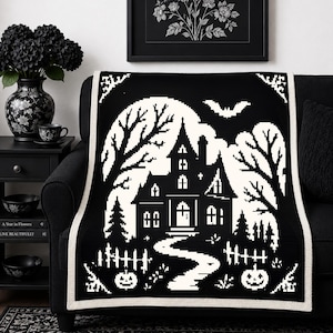 May include: A black and white throw blanket featuring a pixelated haunted house design. The scene includes a large full moon, bare trees, a flying bat, a winding path, a picket fence, and two jack-o'-lanterns, all framed by a solid white border.