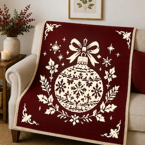 Op de afbeelding: Een dieprode plaid met een crèmekleurige, gepixelde afbeelding van een grote kerstbal met een strik. Het ontwerp bevat omliggende sneeuwvlokken, hulstbladeren en decoratieve hoekaccenten met een crèmekleurige rand.