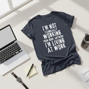 May include: A dark navy blue short-sleeved t-shirt featuring white distressed text that reads: I'M NOT TECHNICALLY WORKING FROM HOME ANYMORE I'M LIVING AT WORK. The shirt has a crew neckline and is displayed flat with a casual, relaxed fit.