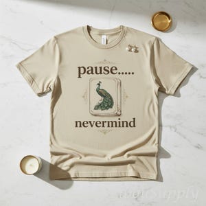 May include: A beige short-sleeve t-shirt featuring the text "pause....." above a graphic of a peacock on a rectangular tile, with the text "nevermind" below. The design is centered on the chest of the shirt in a dark brown font.