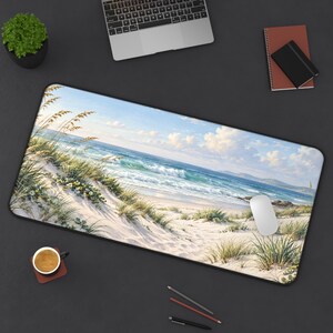 May include: A rectangular desk mat featuring a printed landscape of a sandy beach with tall dune grasses, white flowers, and blue ocean waves under a bright sky. The mat is positioned on a dark surface alongside a laptop, a notebook, a coffee mug, and pens.