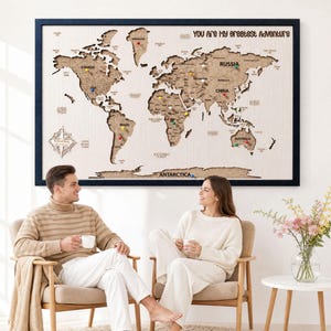 May include: A large rectangular wall map featuring laser-cut wooden continents mounted on a light-colored background with a dark blue frame. The text "YOU ARE MY GREATEST ADVENTURE" is printed at the top, and colorful push pins mark various locations.