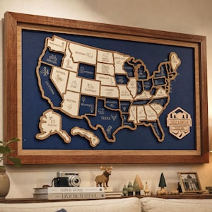 Peut inclure: Une carte en bois encadrée des États-Unis présentant des découpes d'États individuels avec des noms et des icônes gravés, sur un fond bleu foncé. Une plaque hexagonale en bois à droite affiche le texte Lucas & Amelia Travel Map.