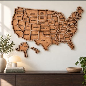 May include: A wall-mounted map of the United States constructed from cork and wood. The map features individual state sections with black text labels for each state name. The design includes separate cutouts for Alaska and Hawaii, showcasing a detailed texture.
