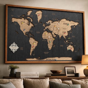 May include: A rectangular world map wall art piece featuring a dark wood-textured background and light-colored cork continents. The map includes a white compass rose with the text The Daniel Family USA Travel Map and colorful push pins.