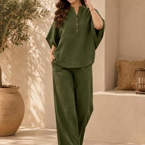 May include: An olive green two-piece set consisting of a short-sleeved top with a mandarin collar, a partial button-down front with four visible buttons, and wide-leg trousers. The garment features a textured fabric and a relaxed, loose fit.