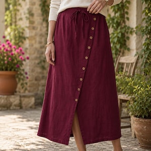 May include: A deep burgundy midi skirt featuring a diagonal button-down front closure with light brown buttons and a drawstring waist. The skirt is paired with a long-sleeved cream-colored top and simple brown flat shoes, creating a cohesive look.
