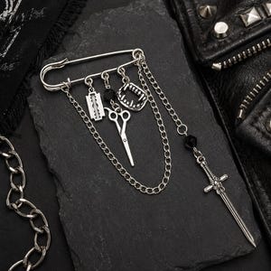 May include: A silver-toned safety pin brooch featuring dangling charms including a razor blade, a pair of scissors, vampire fangs, and a sword. The charms are connected by silver chains and accented with small black beads for a layered look.