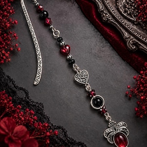 May include: A silver-toned metal bookmark featuring an intricate scrollwork pattern on the hook. The attached chain is adorned with faceted red and black beads, a small silver heart charm, and a larger silver pendant with a red stone.