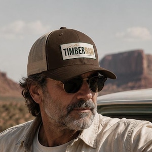 Puede incluir: Una gorra tipo camionero de color marrón y beige con un parche rectangular en la parte frontal que muestra el texto TIMBERRAW en letras de color verde oscuro. La gorra cuenta con un panel frontal estructurado, malla trasera y visera curva.
