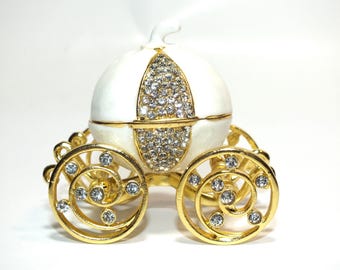 May include: A white porcelain Cinderella carriage trinket box with gold accents and clear crystal embellishments. The carriage has two wheels with intricate designs.