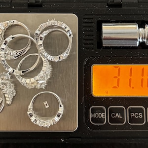 May include: Several silver rings with clear gemstones are arranged on a digital scale. The scale displays a weight of 31.10 grams. The rings vary in design, with some featuring multiple stones and others a single, prominent gem.