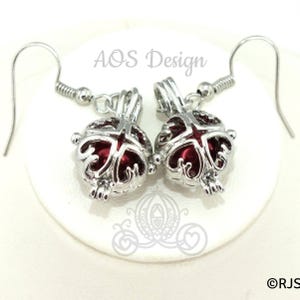 May include: A pair of silver-toned earrings with a decorative design. Each earring features a round, openwork cage with a deep red interior. The earrings have a hook closure and are displayed on a white surface. The text "AOS Design" is visible.