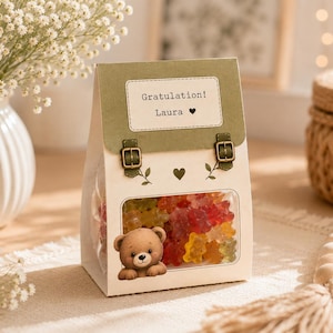 Könnte beinhalten: Eine cremefarbene Geschenktüte aus Papier mit einer grünen Klappe, zwei kleinen Schnallen und einem Etikett mit der Aufschrift Gratulation! Laura und einem Herzsymbol. Die Tüte hat ein Sichtfenster mit bunten Gummibärchen und einem Bärenmotiv.