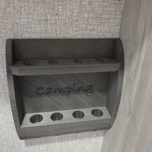 May include: A gray wall-mounted organizer with two horizontal shelves, each featuring four circular cutouts for holding items. The word "camping" is embossed in a cursive font on the back panel between the two shelves.