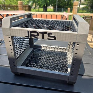 May include: A gray, cube-shaped storage crate featuring a black honeycomb mesh pattern on the top and sides. The frame is constructed with vertical pillars containing small circular holes, and the front panel displays the black text JRTS.