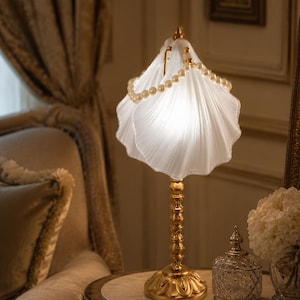 May include: A table lamp featuring a white frosted glass shade shaped like a seashell, adorned with a string of white pearls draped over the top. The lamp has a slender, textured gold-colored metal stand and a decorative, ornate circular base.