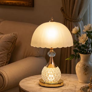 May include: A table lamp featuring a scalloped, frosted glass dome shade, a gold-toned metal neck, and a spherical, textured crystal glass base. The lamp emits a warm, soft glow from within the base and the shade, resting on a circular gold base.