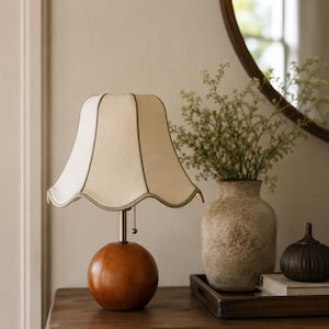 May include: A table lamp featuring a spherical, polished brown wooden base and a cream-coloured fabric lampshade with a scalloped edge and thin piping. A gold-toned metal stem connects the base to the shade, which includes a pull chain switch.