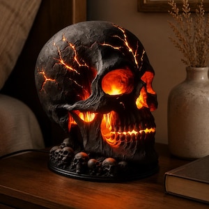 May include: A black, textured skull-shaped lamp featuring glowing orange light emanating from deep cracks across the cranium and through the eye sockets, nose, and teeth. The base is decorated with several smaller, unlit miniature skulls.