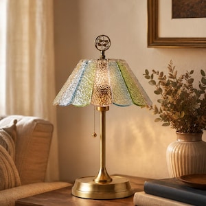 May include: A table lamp with a brass-toned metal base and a scalloped, textured glass shade featuring alternating panels of light blue, green, and amber. A small circular finial with a dragonfly motif sits atop the shade, and a pull chain is attached.