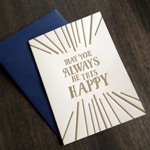 May include: A white card with gold lettering that reads "May you always be this happy". The card has a gold sunburst design.