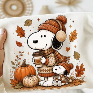 Op de afbeelding: Een crèmekleurig t-shirt met een grafische illustratie van twee witte cartoonhonden die oranje gebreide mutsen en sjaals dragen. De honden houden koffiebekers vast te midden van vallende herfstbladeren en twee oranje pompoenen.