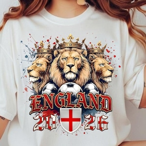 Könnte beinhalten: Ein weißes T-Shirt mit einer Grafik von drei Löwen mit goldenen Kronen über drei Fußbällen. Unter den Löwen steht der Text ENGLAND 2026, ergänzt durch ein Wappen mit einem roten Kreuz auf weißem Hintergrund.