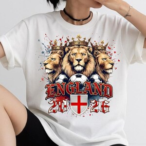 May include: A white t-shirt featuring a graphic of three lions wearing gold crowns, positioned above three soccer balls. Below the lions, the text reads ENGLAND 2026, accompanied by a shield displaying a red cross on a white background.