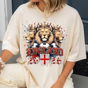 May include: A cream-colored t-shirt featuring a graphic of three lions wearing crowns above soccer balls. The text reads ENGLAND 2026, accompanied by a shield displaying the red cross of Saint George and red and blue paint splatter accents.