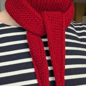 May include: A bright red knitted scarf with a textured garter stitch pattern, tied in a simple knot around the neck. The scarf is worn over a shirt featuring horizontal navy blue and white stripes, creating a clear contrast between the two items.