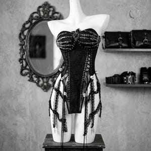 May include: A black strapless corset bodysuit featuring a skeletal hand print across the chest, lace-up side panels, and dangling lace trim accents adorned with small skull beads. The garment is displayed on a white mannequin torso.