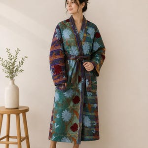 May include: A long-sleeved, calf-length quilted robe featuring a vibrant patchwork design with floral patterns in shades of teal, blue, and deep red. The garment includes a matching fabric tie belt at the waist and a shawl collar, constructed with visible stitching throughout the fabric.