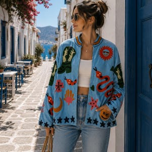 Könnte beinhalten: Eine hellblaue Jacke mit bunten Stickmotiven, darunter ein grüner Cowboystiefel, eine rote Sonne, eine gewundene orangefarbene Schlange, ein rosa Seestern, eine gelbe Mondsichel und schwarze Sterne am Saum, getragen über einem weißen bauchfreien Oberteil.