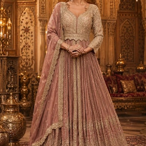 Pode incluir: Um vestido longo até ao chão em rosa seco com mangas compridas, decote em V e um corpete intrincadamente bordado a ouro. A saia é fortemente plissada com uma bainha larga dourada recortada, acompanhada por uma dupatta transparente a condizer.