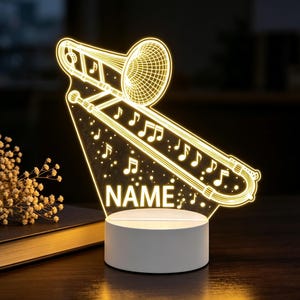 Op de afbeelding: Een oplichtend LED-nachtlampje met een helder acryl paneel gegraveerd met een trombone en muzieknoten. Het ontwerp bevat het woord NAME in hoofdletters. Het paneel is gemonteerd op een ronde witte voet die de afbeelding verlicht.