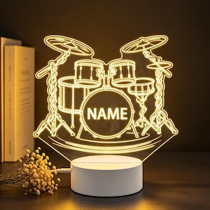 Op de afbeelding: Een oplichtende gele LED-lamp met een lijntekening van een drumstel. Op de centrale basdrum staat het woord NAME in dikke hoofdletters. De lamp is gemonteerd op een ronde witte voet en verspreidt een warm licht in een donkere omgeving.