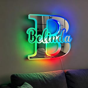 Op de afbeelding: Een grote, aan de muur gemonteerde spiegel in de vorm van de letter B, voorzien van een levendige, meerkleurige LED-rand die door regenboogtinten verloopt. De naam Belinda staat in cursief schrift op het spiegeloppervlak.