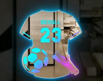 May include: A jersey-shaped wall mirror with a glowing blue LED border. The mirror features the text BESSLL above the number 23, a soccer ball graphic, and a soccer cleat graphic, all rendered in a vibrant blue and purple gradient color scheme.